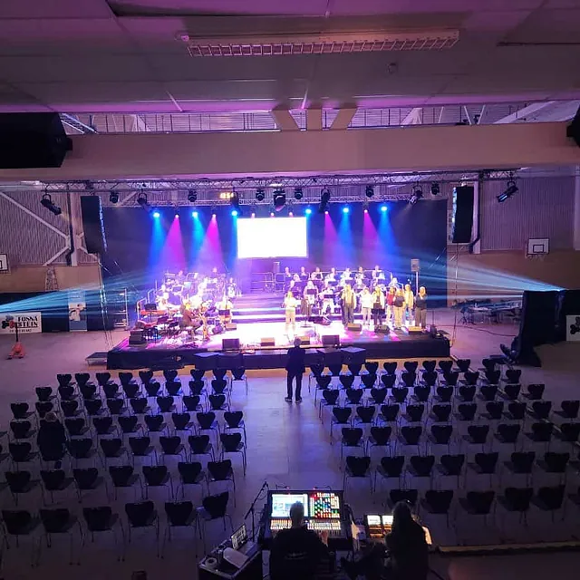 Stor innendørsscene med kor og musikere under konsert i idrettshall.