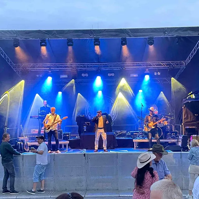 Band spiller på utendørsscene med blå og gule lys, mens folk danser foran.